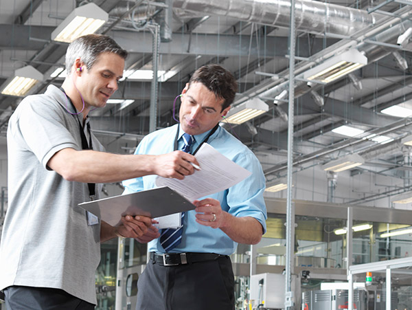 two people in an industrial or office setting reviewing documents on a clipboard, with exposed ductwork and equipment in the background